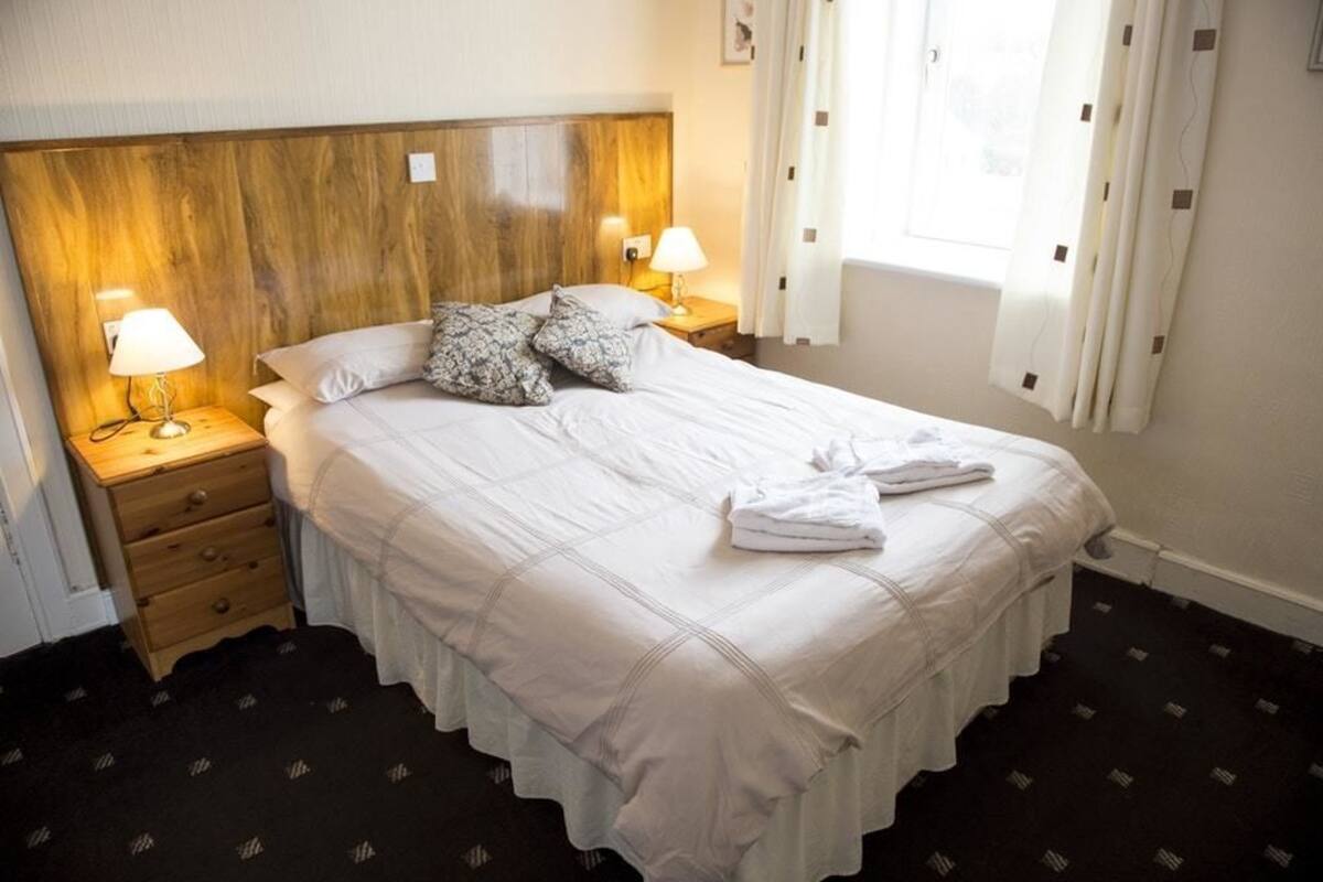 Hotellihuone, jossa on suuri sänky, kaksi yöpöytää valaisimineen, puinen sängynpääty ja ikkuna, jossa on verhot.