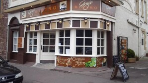 Un bâtiment d’angle, avec une enseigne indiquant « Greg’s Bistro », et une grande vitrine présentant une offre de cocktails à partir de 17 heures. Une autre enseigne, au-dessus de l’entrée, indique qu’il s’agit d’un hôtel.