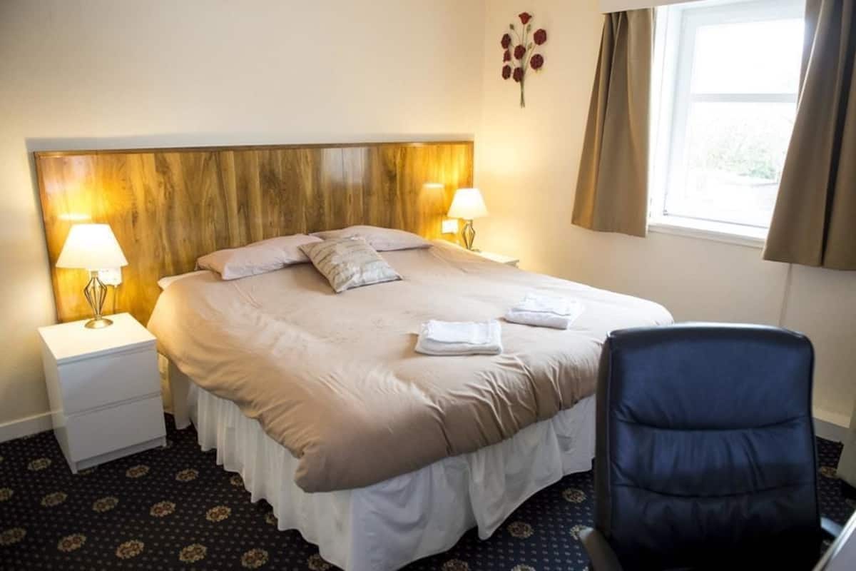 Hotellihuone, jossa on suuri sänky, kaksi yöpöytävalaisinta, tuoli ja ikkuna, jossa on verhot.