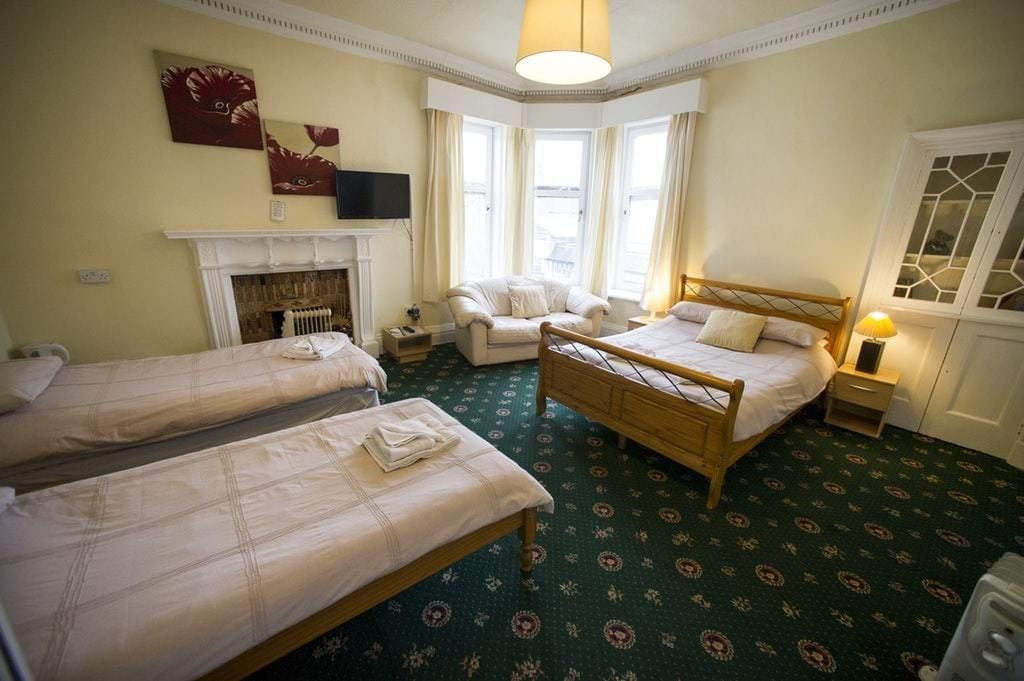 Een hotelkamer met twee enkele bedden, een zithoek met een fauteuil, een open haard en een groot raam met gordijnen.