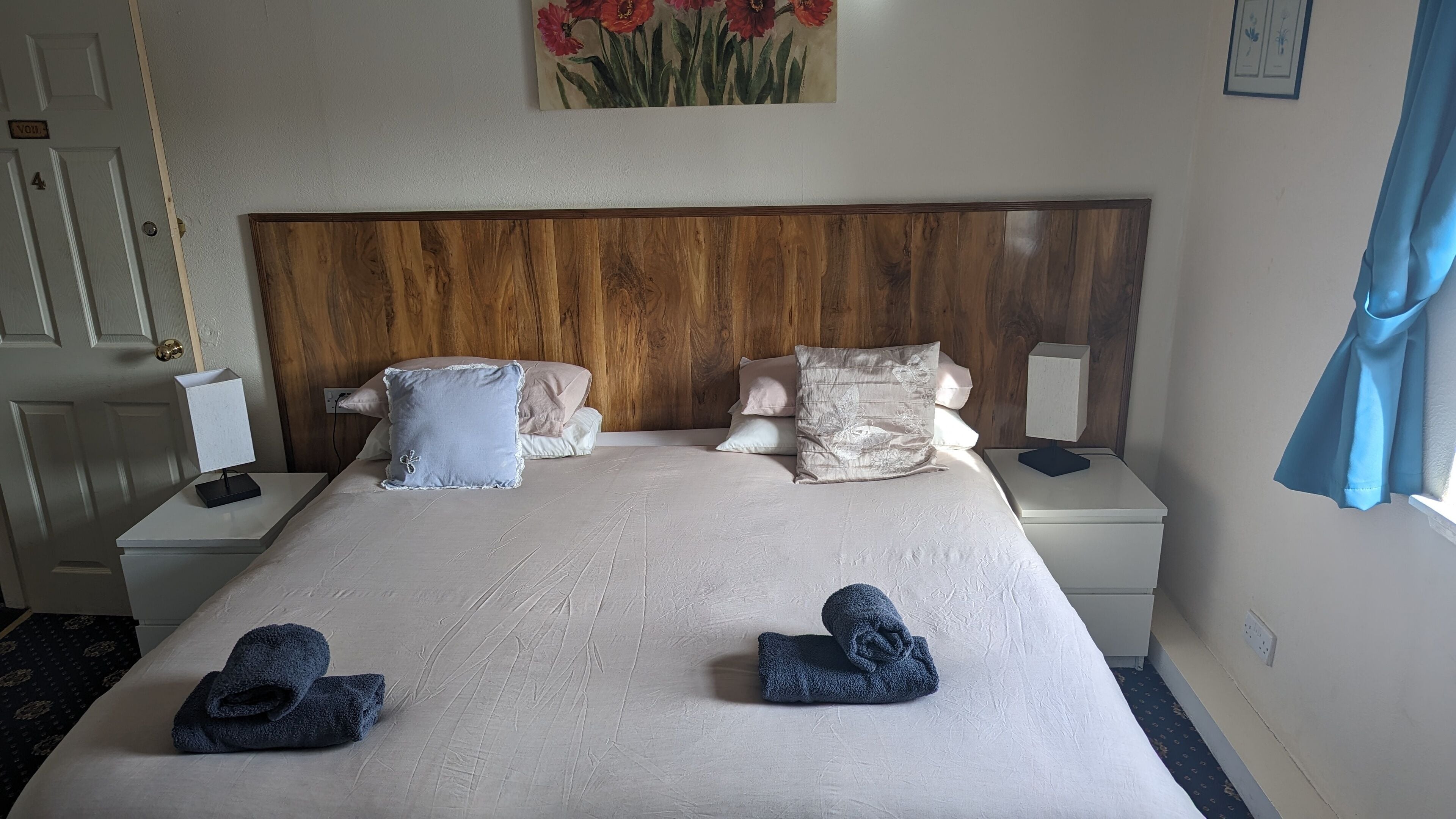 Uma cama bem arrumada com almofadas e toalhas, cabeceira de madeira, mesas de cabeceira com abajures e um quadro na parede.