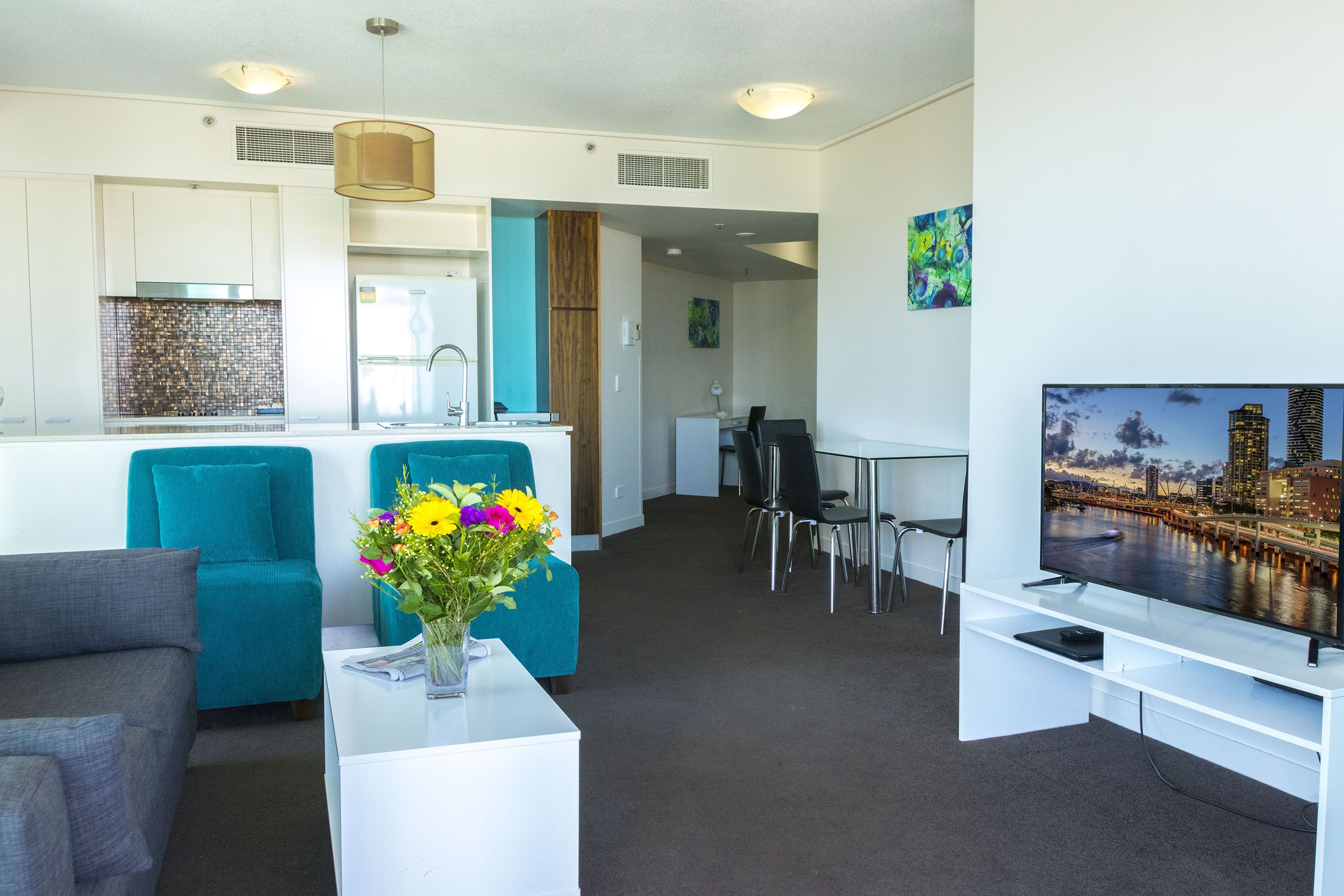 Een moderne woonkamer met teal banken, een witte salontafel met een bloevas, een flatscreen televisie en uitzicht op een stadslandschap.