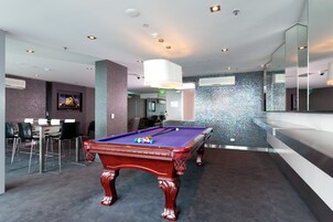 A pool table in a modern room with a carpeted floor, a dining area with chairs, and a wall with a mosaic pattern.