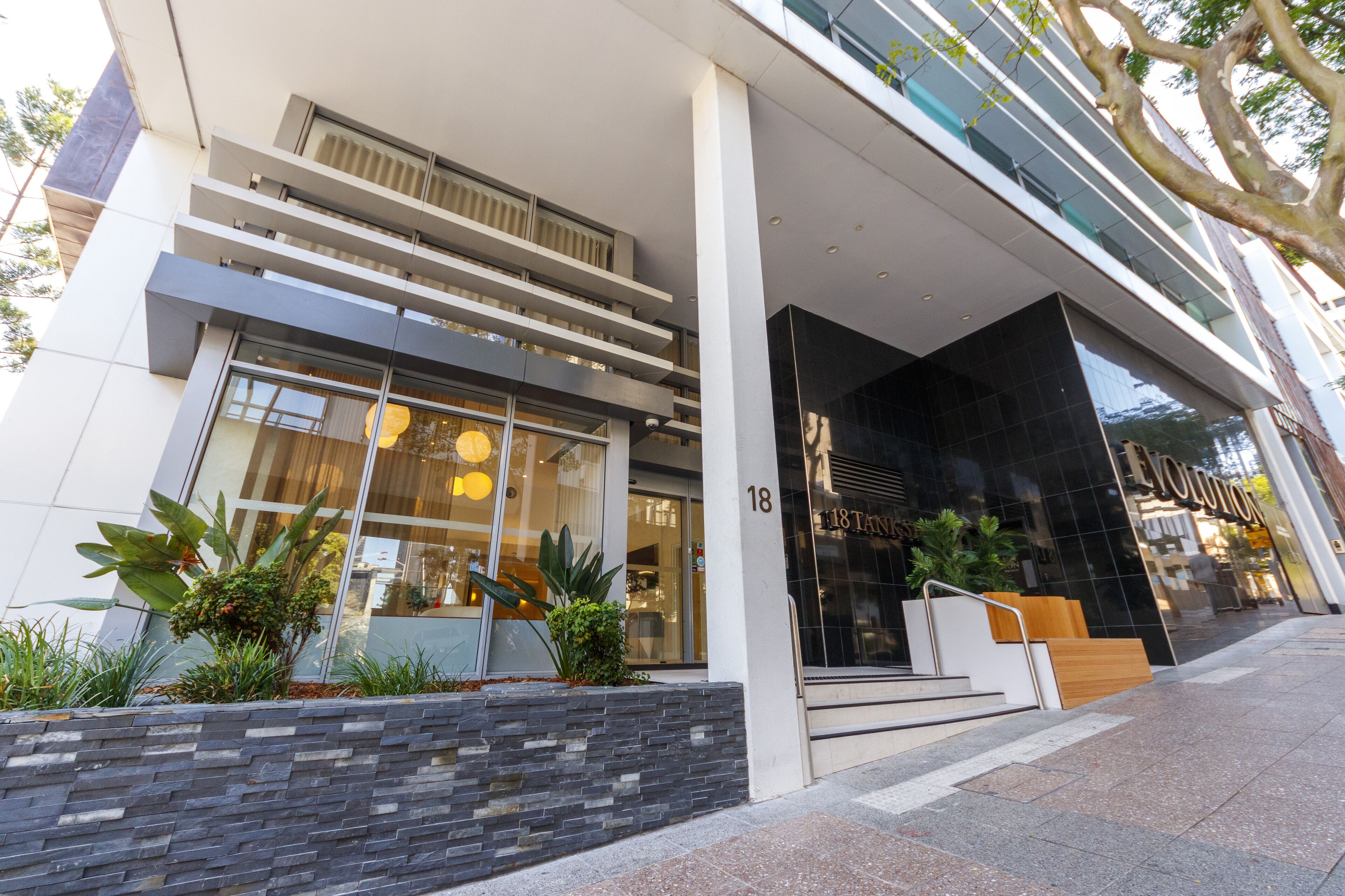 Un bâtiment moderne doté d’une façade en verre, d’une entrée bien visible avec des marches et du chiffre « 18 » peint sur le mur.