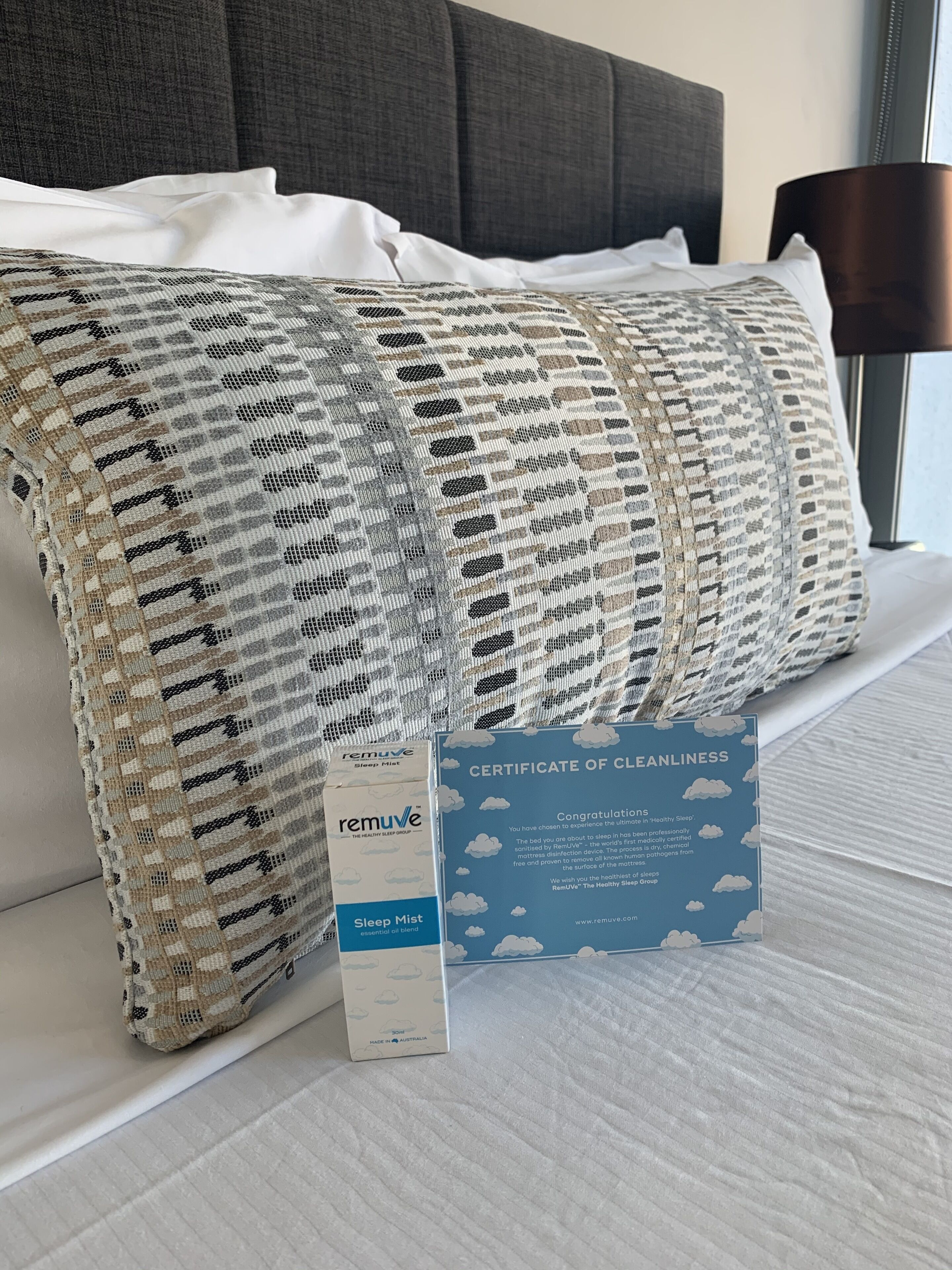 Una cama bien hecha con un edredón y almohadas estampadas, un certificado de limpieza y un atomizador para dormir.