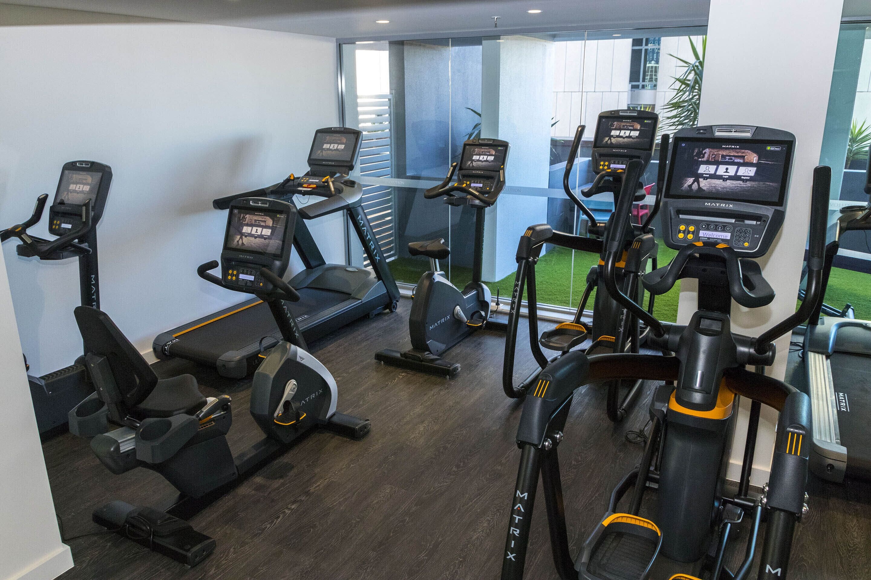 Un gimnasio con varias bicicletas estáticas y cintas de correr, suelo de madera y vistas a un espacio exterior verde a través de amplias ventanas.
