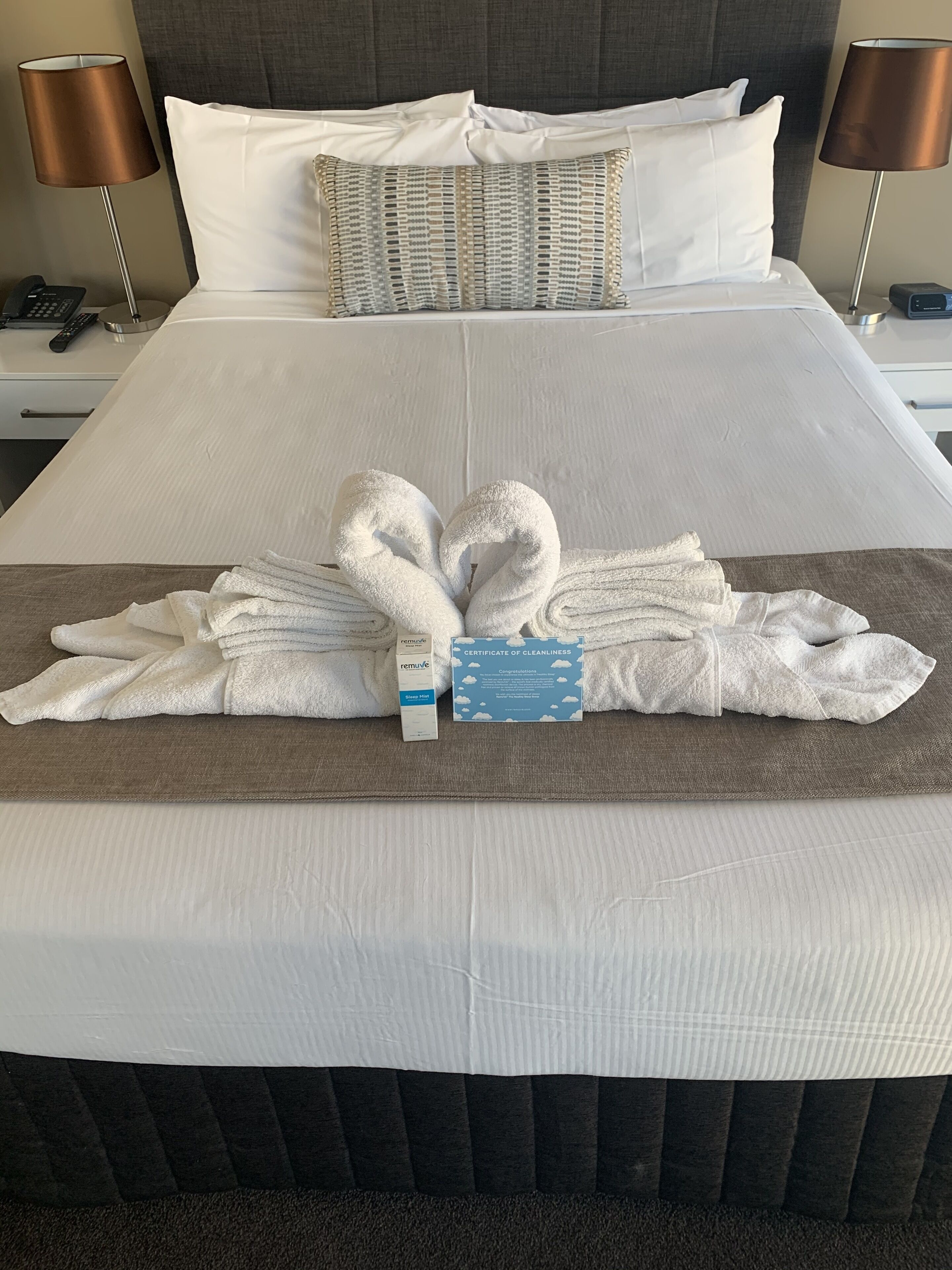 A neatly made bed with a towel arrangement in the shape of a heart, a certificate of cleanliness, and a remote control on the nightstand.