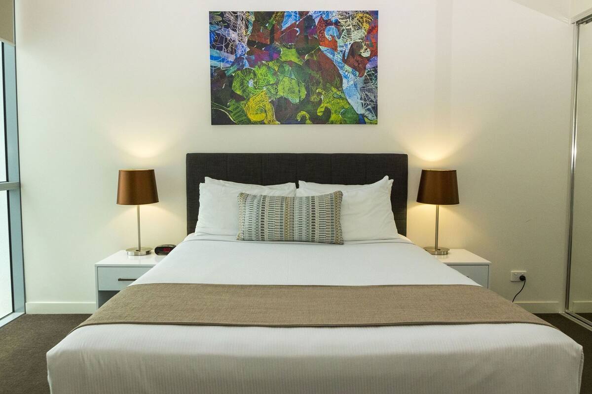 Un lit bien fait, avec du linge de lit blanc, deux lampes de chevet, une grande peinture abstraite au-dessus du lit et une fenêtre sur le côté gauche.
