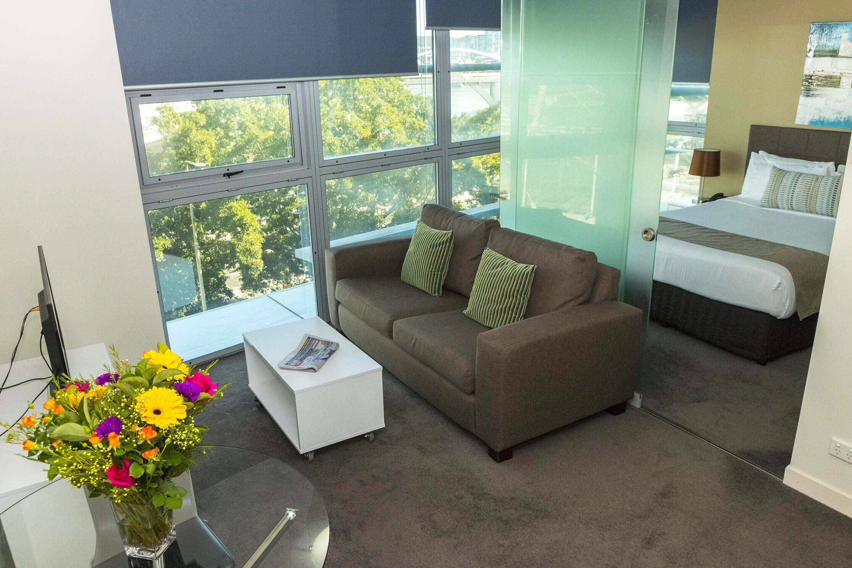 A modern hotel room with a large window, a brown sofa, a white side table, and a bed with a headboard.