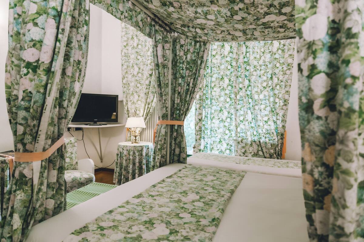 Una habitación de hotel con cortinas y ropa de cama estampada con motivos florales, un escritorio con televisor y una mesita de noche con lámpara.