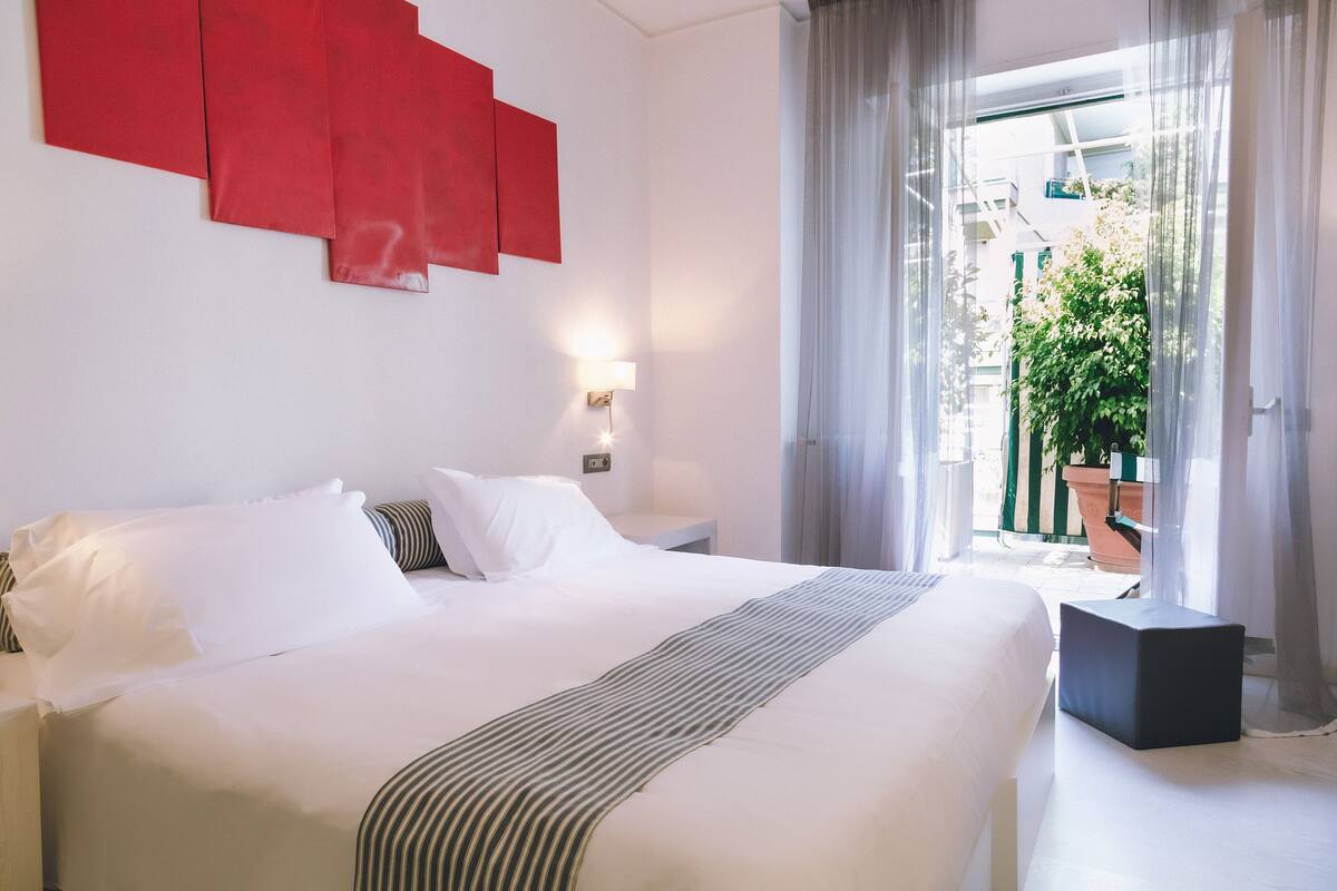 Habitación de hotel con una cama grande, una mesita de noche y una pared de acento roja, con vistas a un balcón con plantas.