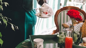 Une personne portant un gilet vert et une chemise blanche, qui porte un plateau avec une théière, des tasses et une rose.