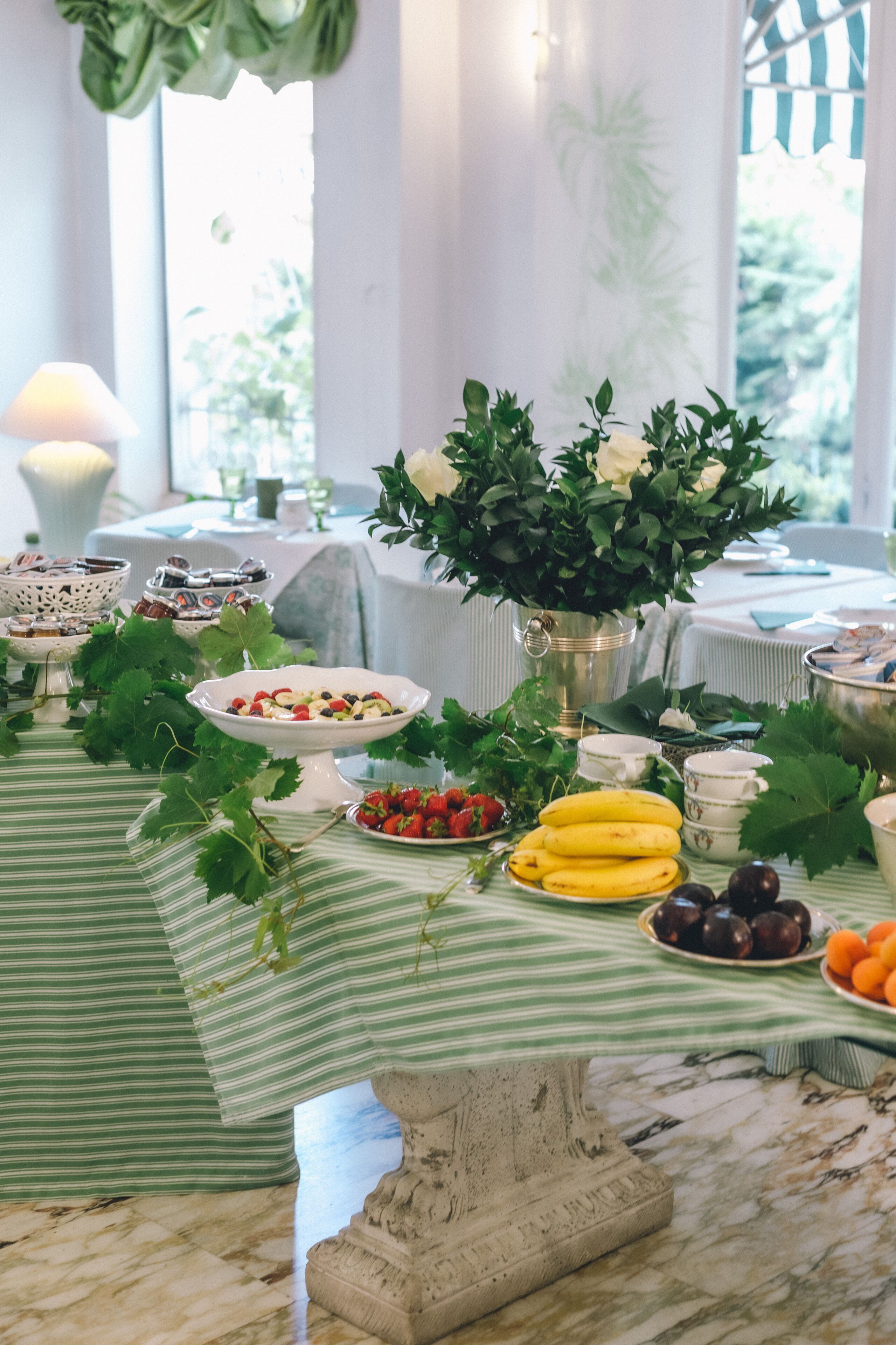A table set with various fruits and a vase of flowers, with a striped tablecloth and a marble floor.