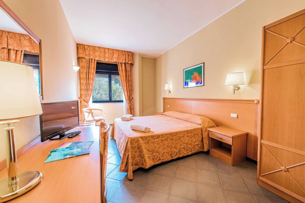 Une chambre d’hôtel avec un lit, une table de chevet, une lampe, une chaise et une fenêtre avec des rideaux.