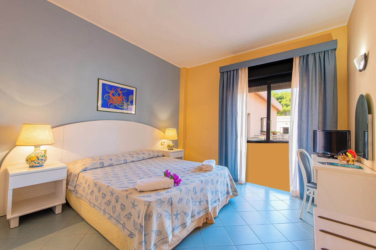 Habitación de hotel con cama, dos mesitas de noche con lámparas, televisor, escritorio con silla y ventana con cortinas.