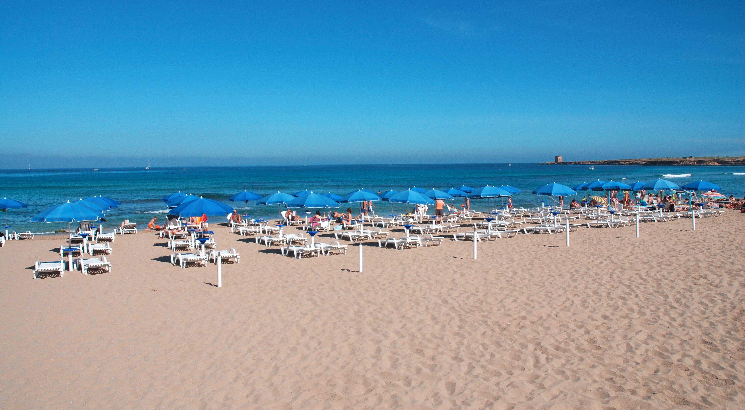 A beach with rows of white sun loungers and blue umbrellas.