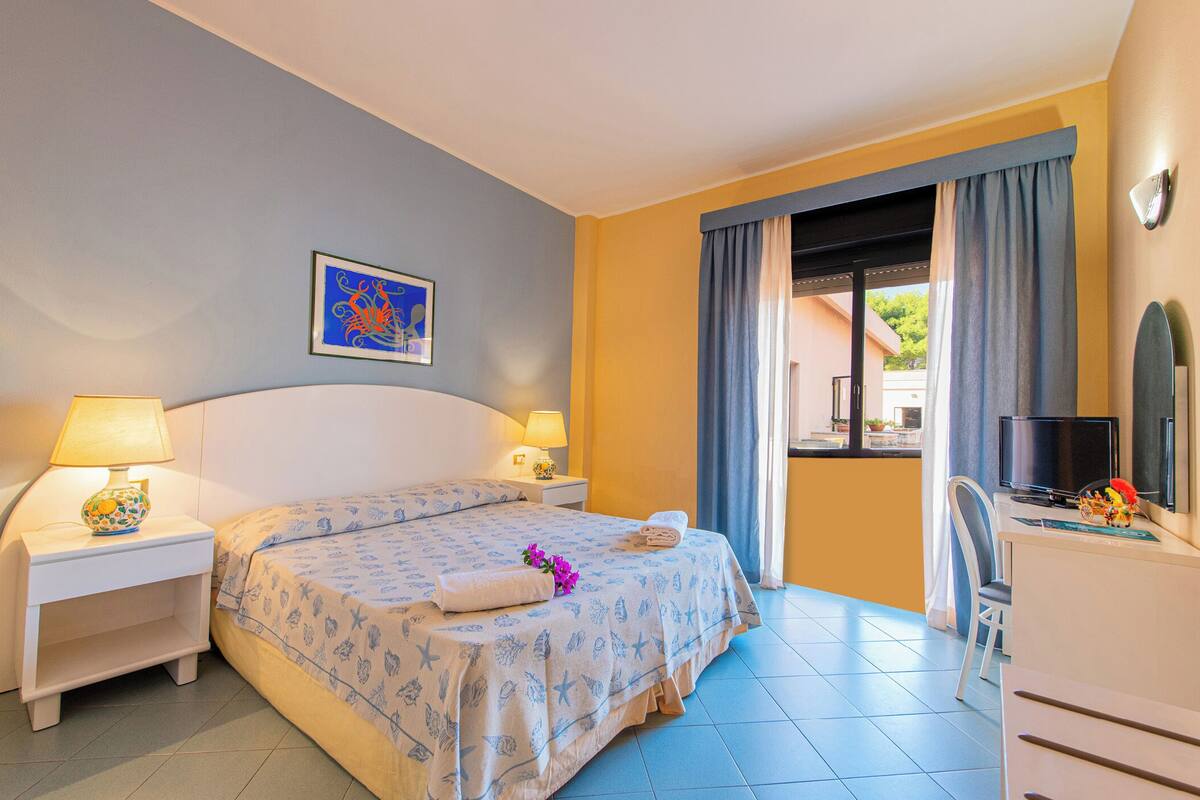 Une chambre d’hôtel comprenant un lit, deux tables de chevet avec des lampes, une télévision, un bureau avec une chaise et une fenêtre avec des rideaux.