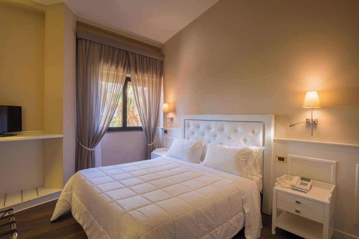 Une chambre d’hôtel avec un lit, une table de chevet avec un téléphone, un téléviseur posé sur une étagère et une fenêtre avec des rideaux.