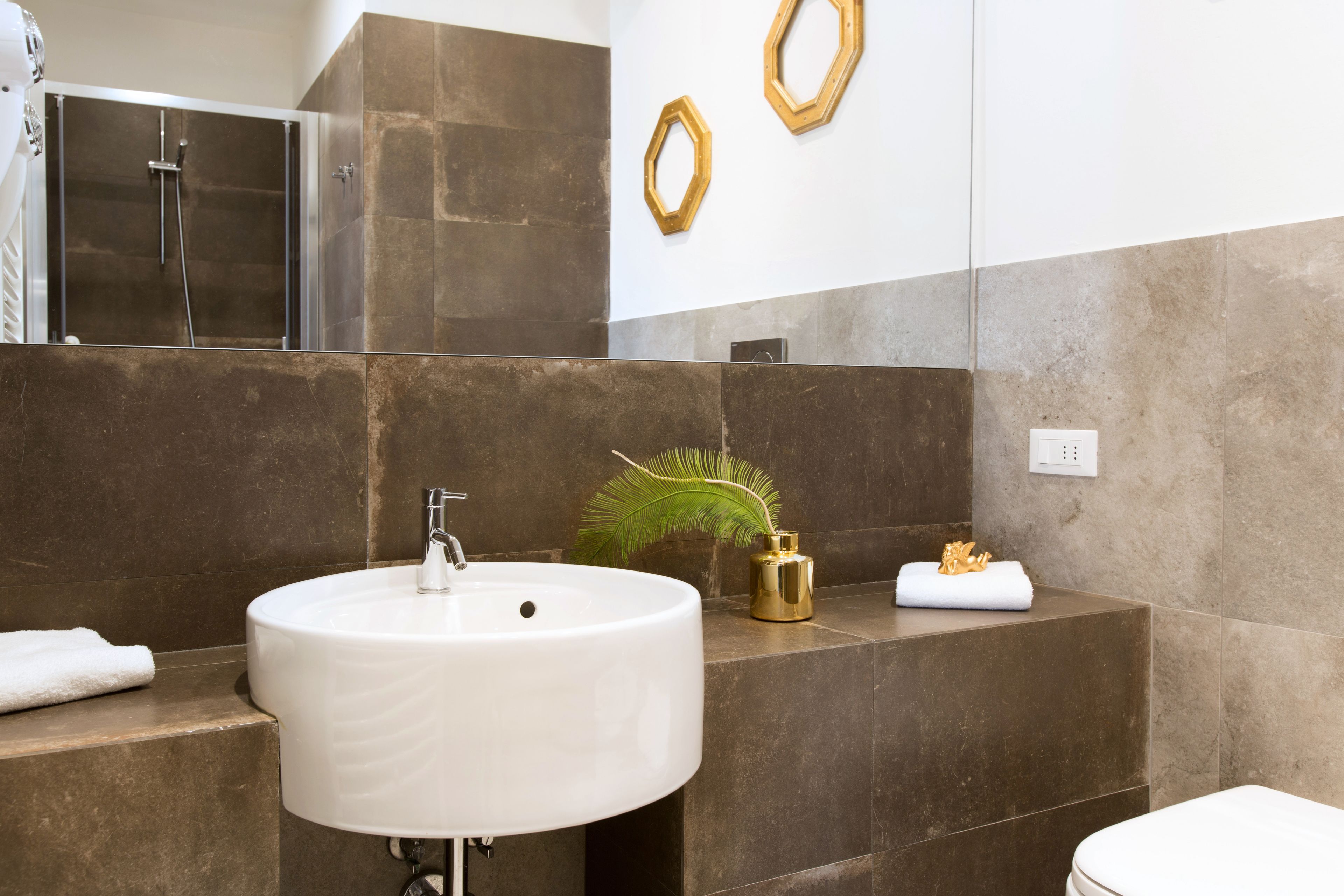 Un baño moderno con un lavamanos grande de color blanco, una ducha y un inodoro. Hay detalles decorativos dorados en las paredes y una planta pequeña en la encimera.