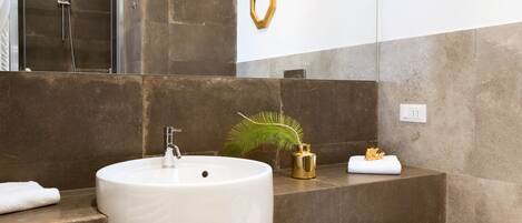 Un baño moderno con un lavamanos grande de color blanco, una ducha y un inodoro. Hay detalles decorativos dorados en las paredes y una planta pequeña en la encimera.