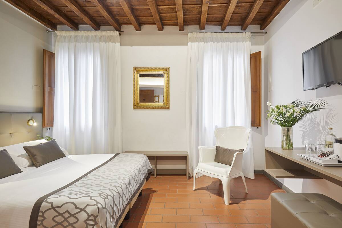A hotel room with a bed, a chair, a TV, and a small table with a vase of flowers.
