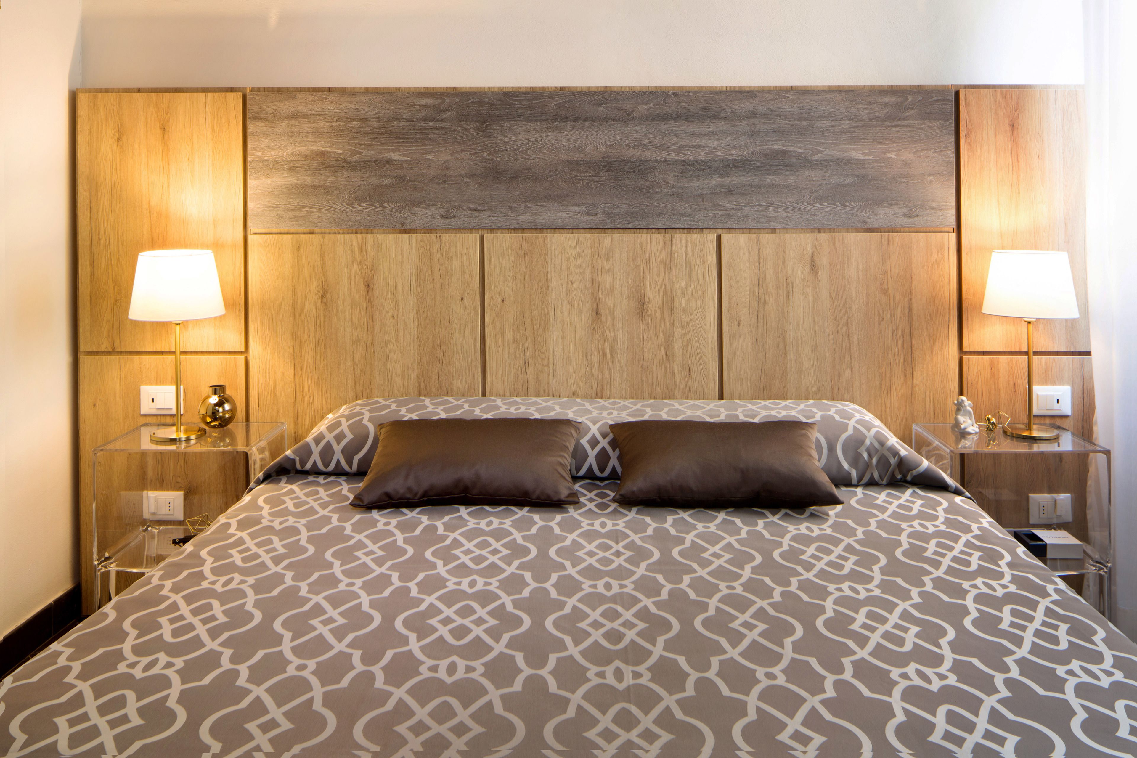A hotel room with a wooden headboard, two bedside lamps, a patterned bedspread, and a nightstand with a lamp and a small statue.