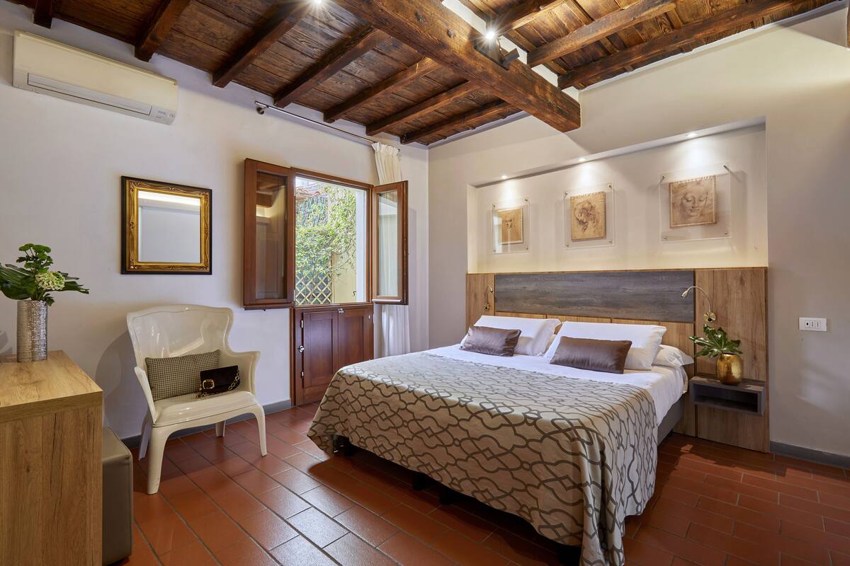 A bedroom with a large bed, wooden ceiling beams, a chair, a bedside table, and artwork on the wall.