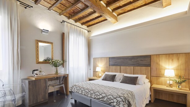 A modern bedroom with a wooden ceiling, a large bed, bedside tables with lamps, a desk with a chair, and a mirror on the wall. - Sette Angeli Rooms (Florence)