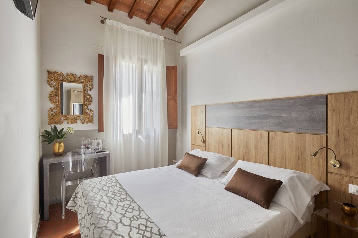A hotel room with a large bed, a wooden headboard, a desk with a chair, a mirror, and a window with curtains.