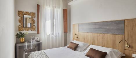 A hotel room with a large bed, a wooden headboard, a desk with a chair, a mirror, and a window with curtains.