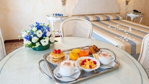 Bandeja de desayuno con tortitas, café, cereales y fruta sobre una mesa de vidrio en una habitación de hotel.
