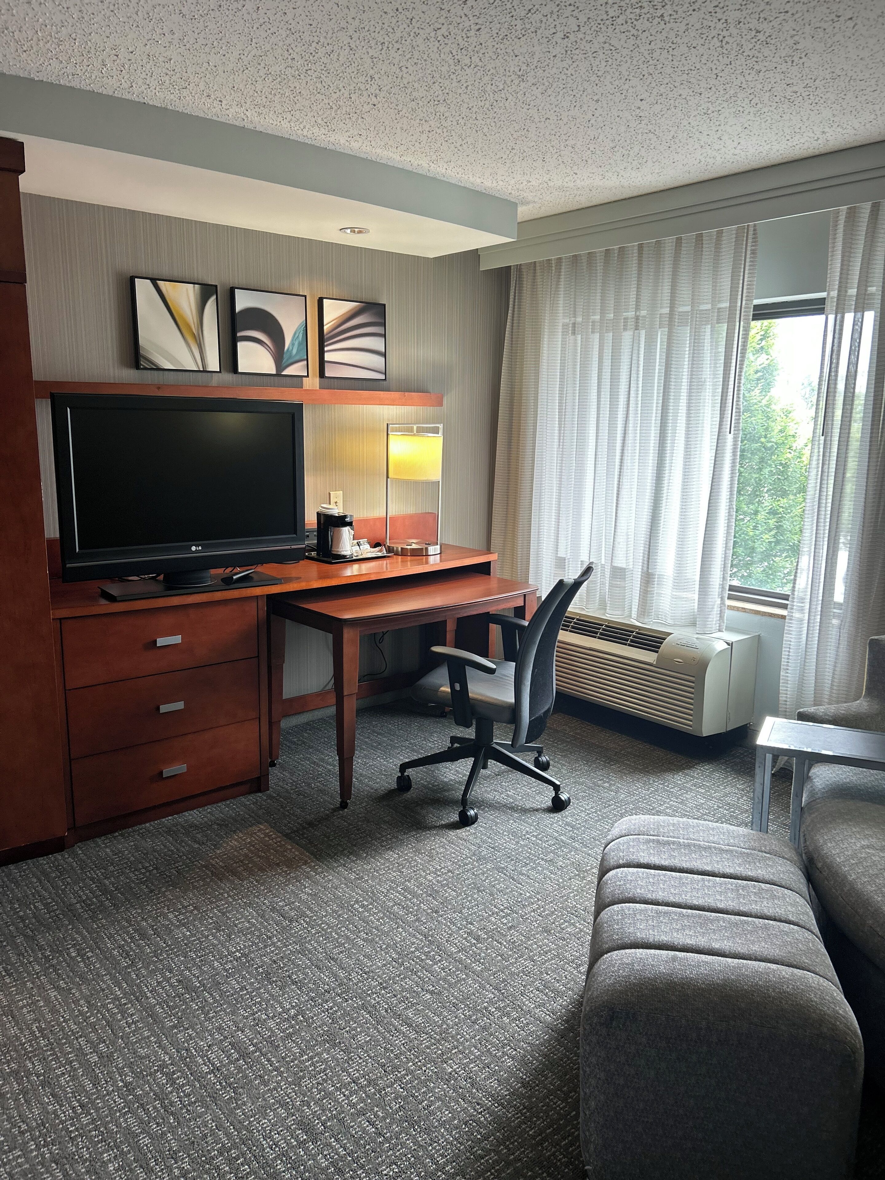 A hotel room with a wooden desk, a flat-screen TV, a lamp, a chair, an air conditioner, and a window with curtains.