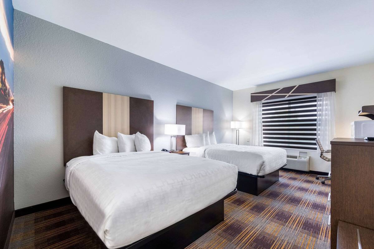 A hotel room with two beds, a desk, a chair, a lamp, and a window with blinds.