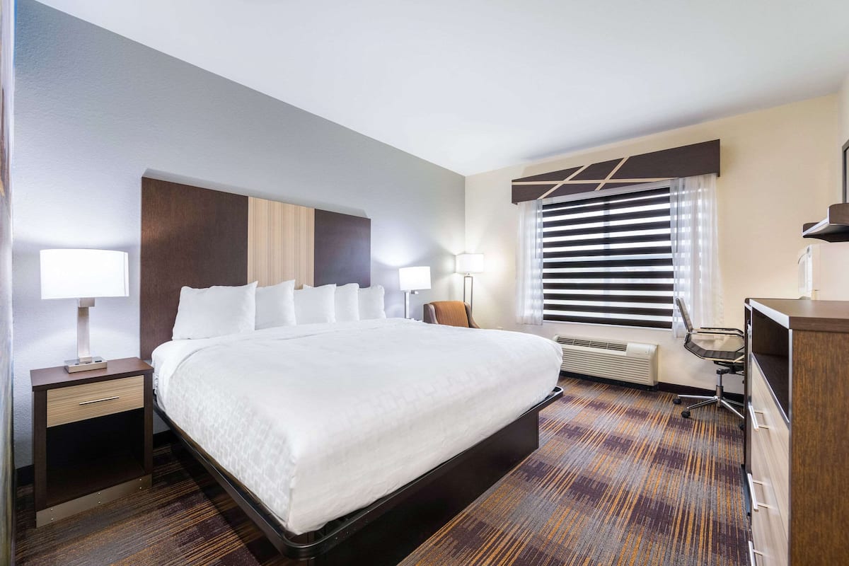 A hotel room with a large bed, bedside tables with lamps, a desk with a chair, a window with blinds, and a built-in heater.