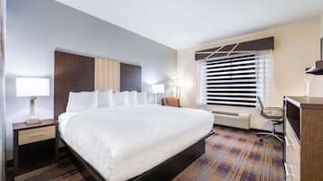 Ein Hotelzimmer mit einem großen Bett, Nachttischen mit Lampen, einem Schreibtisch mit Stuhl, einem Fenster mit Jalousien und einer fest installierten Heizung.