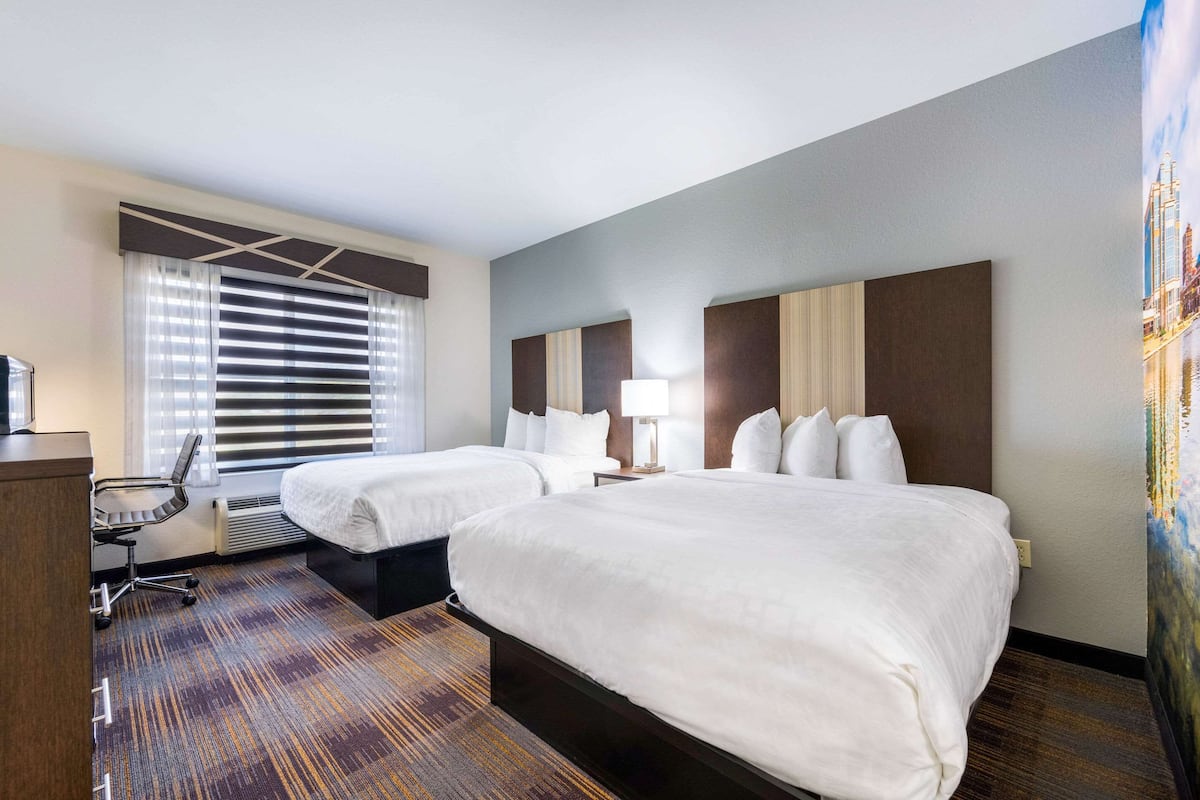 A hotel room with two beds, a desk, a chair, and a large window with blinds.