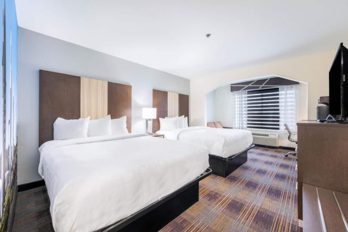 A hotel room with two beds, a desk, a chair, and a large window with blinds.