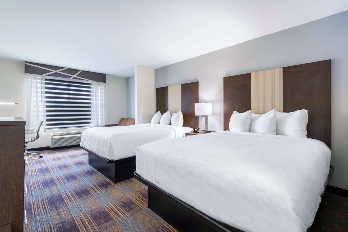 A hotel room with two beds, a desk, a chair, and a large window with blinds.