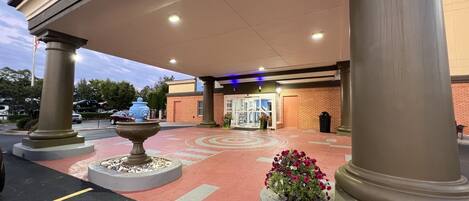 A building entrance with a covered walkway, columns, a fountain, and a flower bed.