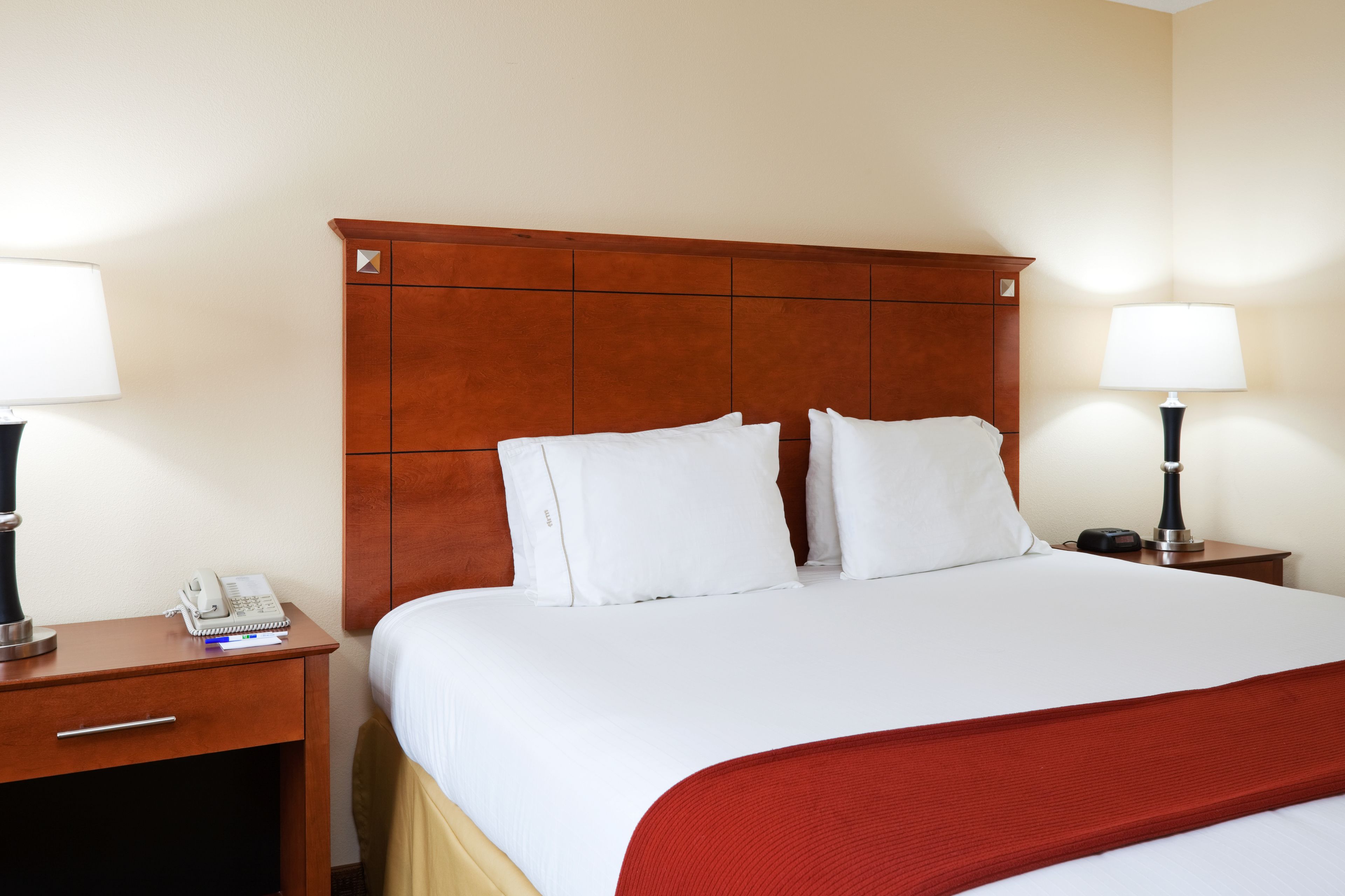A hotel room with a wooden headboard, white bedding, a red throw blanket, two bedside tables with lamps, and a phone on one table.