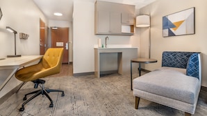 A hotel room with a desk, a yellow chair, a gray sofa, a small round table, a sink, and a mirror.