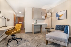 Una habitación de hotel con un escritorio, una silla amarilla, un sofá gris, una mesita redonda pequeña, un lavabo y un espejo.