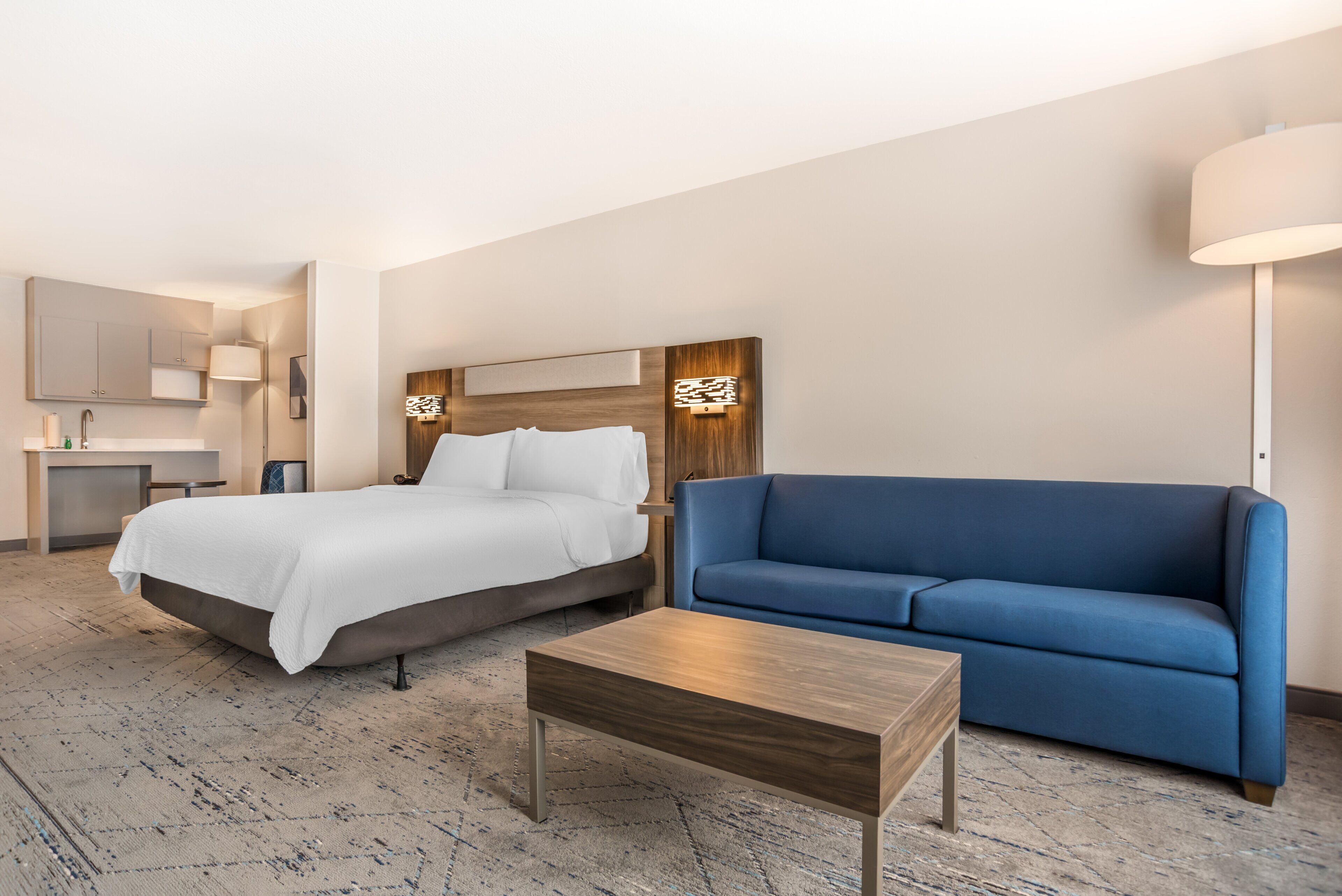 Una habitación de hotel con una cama, un sofá azul, una mesa de centro de madera, un escritorio con una silla y una lámpara.