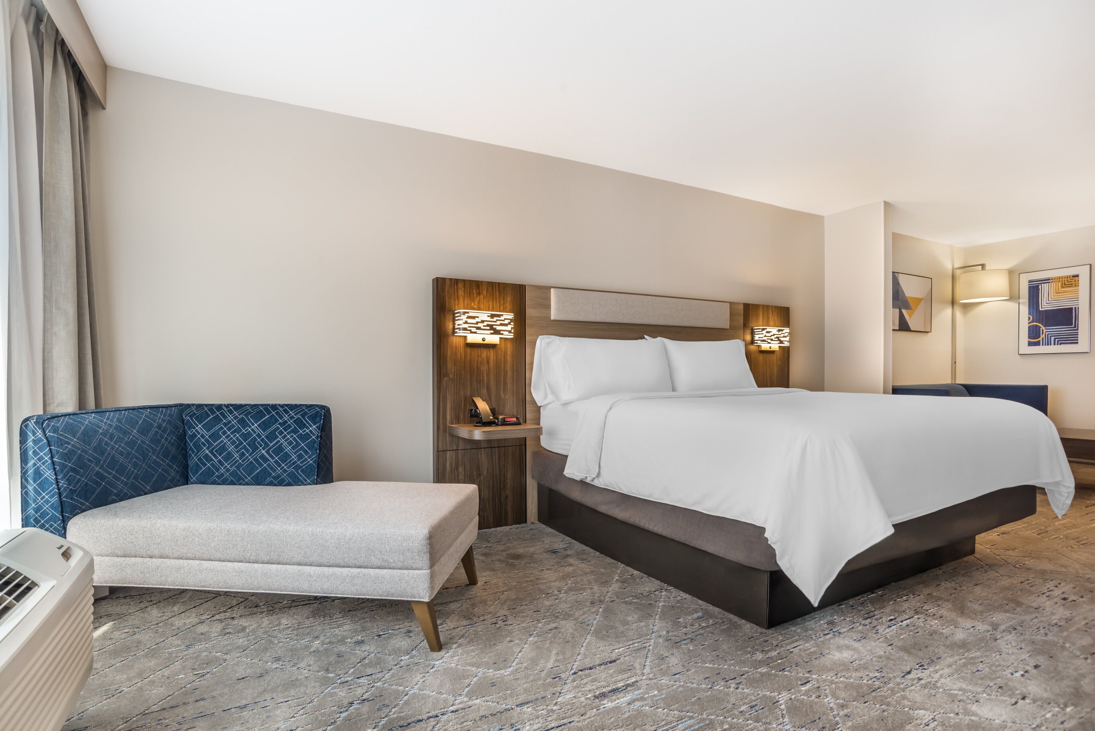 A hotel room with a bed, a seating area, a bedside table, and a wall-mounted light fixture.
