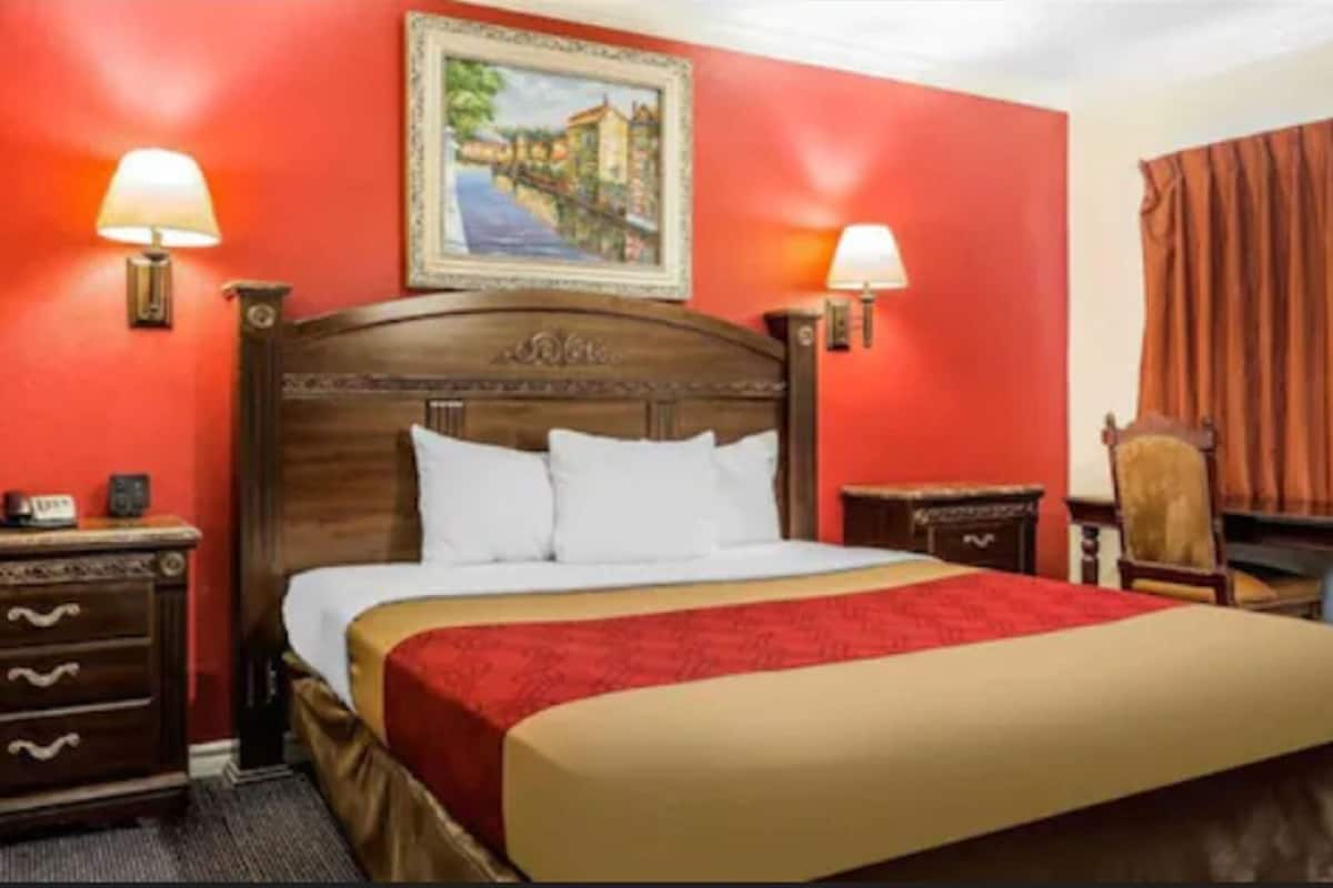 Habitación de hotel con cama de madera, dos mesitas de noche, una silla, un cuadro en la pared y una cortina roja.