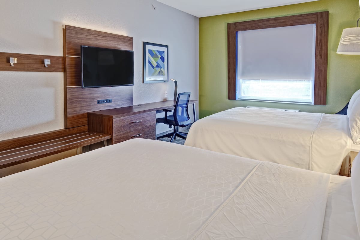 Ein Hotelzimmer mit zwei Betten, einem Schreibtisch mit Stuhl, einem Flachbildfernseher und einem Fenster mit Jalousien.