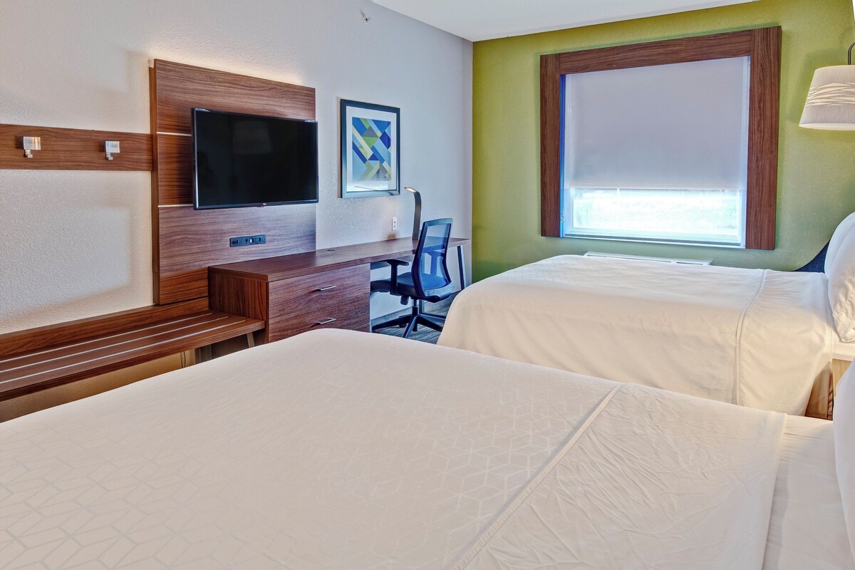 Une chambre d’hôtel avec deux lits, un bureau avec une chaise, un téléviseur à écran plat et une fenêtre avec des stores.