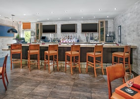 Zona de bar moderna con una barra larga, varios taburetes y un área de estar con una mesa y sillas.
