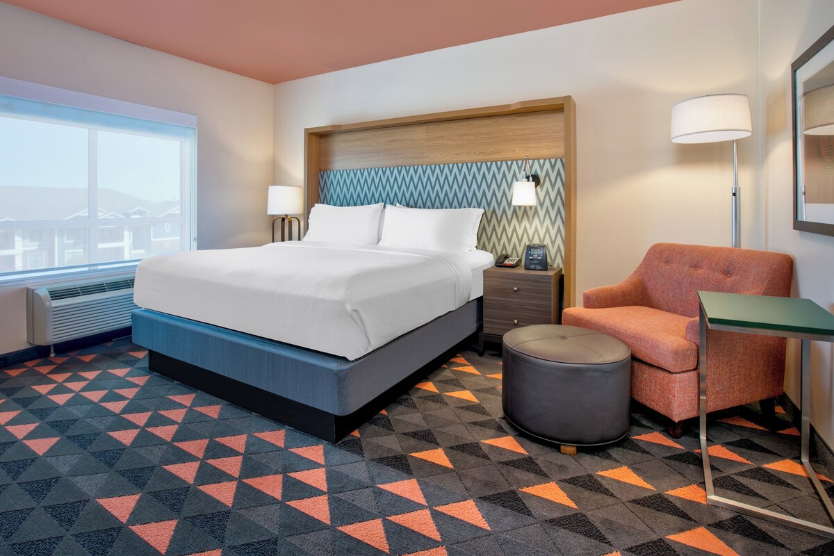 A hotel room with a bed, a chair, a table, a lamp, and a view of buildings outside the window.