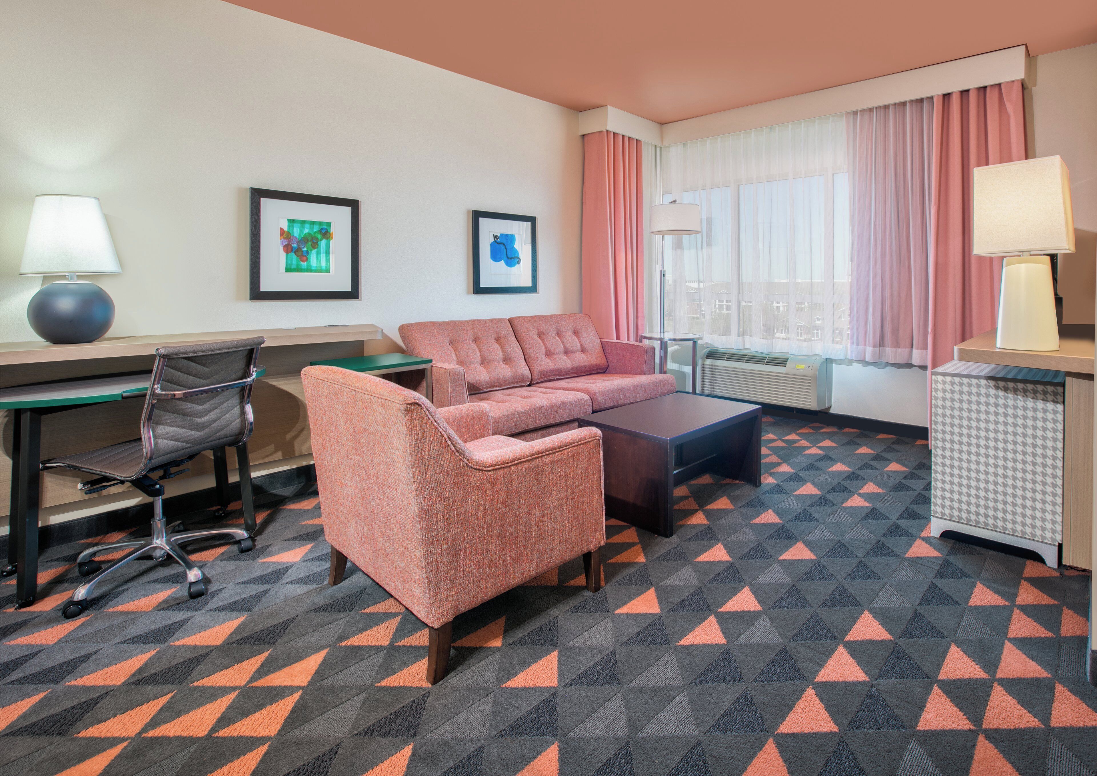 A hotel room with a sofa, armchair, desk, and a patterned carpet.